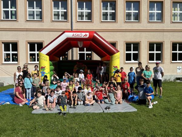 Kinder und Lehrerinnen sitzen vor einer Hüpfburg. Die Sonne scheint. 
